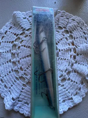 Sale Photo Thumbnail #341: A vintage Rapala Floating balsa wood fishing lure, hand-tuned and tank-tested. Features a blue and silver/white color scheme with three treble hooks. The lure is housed in its original clear plastic slide-top box with Finnish text ('KÄSINTEHTY JA KOETELT