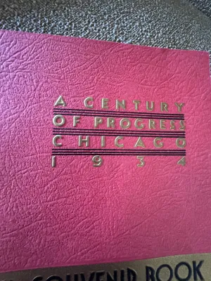 Sale Photo Thumbnail #158: An authentic 1934 souvenir book from the Chicago World's Fair, titled 'A Century of Progress'. This spiral-bound booklet features a textured pink cover with gold-embossed lettering and a central gold seal. Inside, it contains black-and-white photographic 