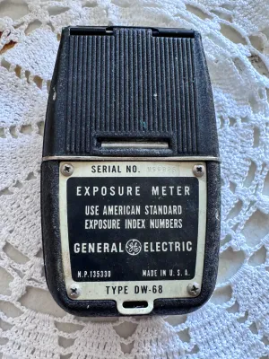 Sale Photo Thumbnail #309: A vintage General Electric Type DW-68 light exposure meter. This classic photographic tool features a black textured body with a dual-dial calculator on the front and a footcandle light scale. The back plate displays the serial number H66825 and is marked