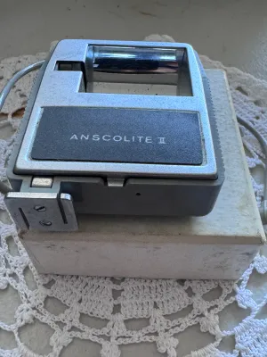 Sale Photo Thumbnail #316: Vintage Anscolite II camera flash unit manufactured by Ansco. Features a grey plastic housing with a built-in exposure guide dial on the back, a red test button, and an integrated PC sync cord. Marked 'Made in Japan' on the back casing. Includes the origi