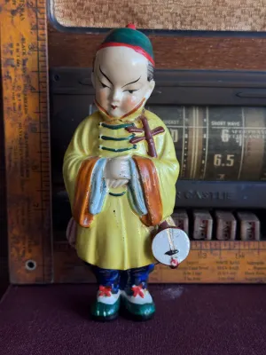 Sale Photo Thumbnail #194: A vintage hand-painted ceramic figurine depicting an Asian boy in traditional yellow and blue attire holding a banjo-like instrument. The base is stamped with a red maker's mark and reads 'Made in Occupied Japan', dating this piece to the post-WWII era (1