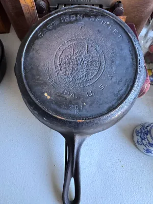 Sale Photo Thumbnail #268: A vintage Griswold No. 7 cast iron skillet, marked with the classic Griswold logo, 'ERIE PA., U.S.A.', and model number '701 E'. Features dual pour spouts and a teardrop handle. The skillet shows signs of age and use, including surface buildup, minor rust