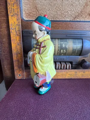 Sale Photo Thumbnail #195: A vintage hand-painted ceramic figurine depicting an Asian boy in traditional yellow and blue attire holding a banjo-like instrument. The base is stamped with a red maker's mark and reads 'Made in Occupied Japan', dating this piece to the post-WWII era (1