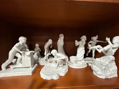 Sale Photo Thumbnail #10: Collection of Norman Rockwell white bisque porcelain figurines by Gorham, depicting various nostalgic scenes of childhood and Americana on a wooden shelf.
