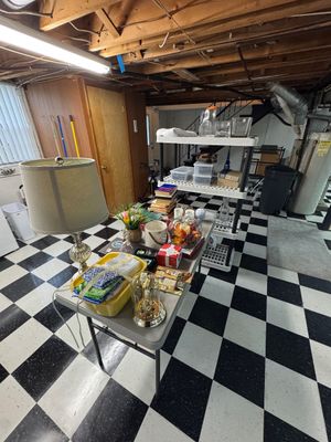 Sale Photo Thumbnail #31: Assorted household items displayed on a folding table and plastic shelving unit, including a table lamp, books, glassware, and decorative pieces in a basement setting.