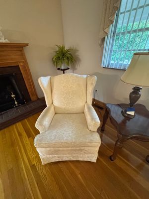 Sale Photo Thumbnail #8: Traditional wingback armchair upholstered in a cream-colored floral damask fabric, shown alongside a dark wood side table and lamp.