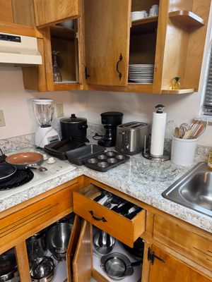 Sale Photo Thumbnail #13: Assorted kitchenware and small appliances, including an Oster blender, a drip coffee maker, a stainless steel toaster, baking pans, and various pots and pans stored in wooden cabinets.