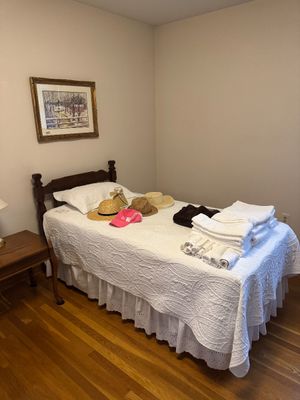 Sale Photo Thumbnail #21: Twin-size wooden bed frame with a white quilted coverlet, featuring an assortment of straw hats, a pink visor, and folded white towels displayed on top.