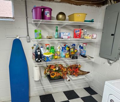 Sale Photo Thumbnail #32: Four-tier white wire shelving unit holding various household cleaning supplies, laundry detergents, an iron, and autumn floral arrangements, shown alongside a blue ironing board.