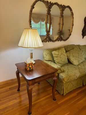 Sale Photo Thumbnail #2: Traditional wooden end table with cabriole legs, paired with a brass and crystal table lamp featuring a pleated shade, set alongside a green damask sofa and an ornate triple-oval gold wall mirror.