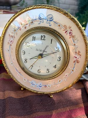 Sale Photo Thumbnail #52: Vintage Saks Fifth Avenue table clock featuring a circular brass frame, hand-painted floral and bow details on a fabric-textured face, and elegant Arabic numerals.