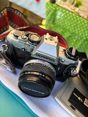 Sale Photo Thumbnail #74: Vintage Olympus OM10 35mm SLR film camera with a 50mm lens, lens cap, and patterned neck strap.