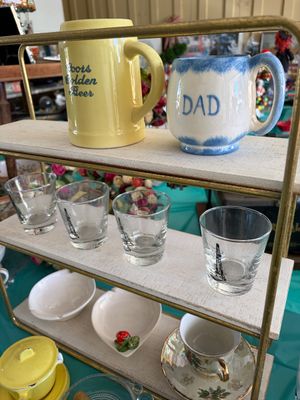 Sale Photo Thumbnail #25: Assorted vintage glassware and ceramics, including a yellow Coors Golden Beer mug, a blue and white "Dad" mug, and four shot glasses featuring oil derrick graphics, displayed on a three-tiered shelf.