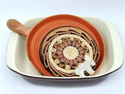 Assorted kitchenware and decor, including a ceramic baking dish, a terracotta pan with a sun-embossed handle, a woven straw trivet, and a...