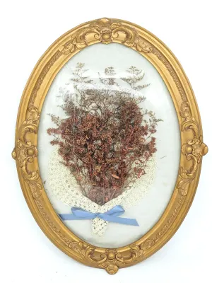 Vintage framed dried flower arrangement under convex glass, featuring a lace wrap and blue ribbon in an ornate oval gilt frame.