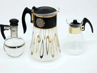 Mid-century modern glass beverage set including a large carafe with gold and black atomic designs, a smaller glass server with a gold...