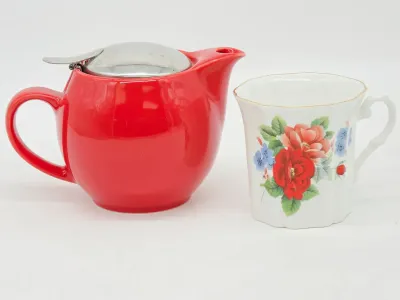 Modern red ceramic teapot with a hinged metal lid, paired with a vintage-style white porcelain teacup featuring a floral rose design.