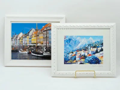 Two framed art prints featuring European landscapes, including a colorful harbor scene and a mountain village, both housed in white frames.