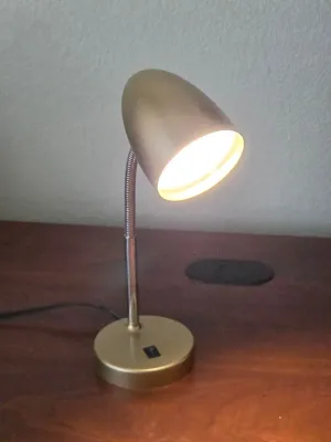 Modern gold-finished desk lamp featuring an adjustable gooseneck arm and a bullet-shaped shade, shown illuminated on a wooden surface.