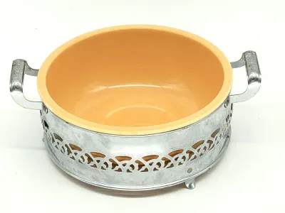 Vintage yellow ceramic casserole dish nestled in a footed chrome carrier with ornate openwork detailing and side handles.