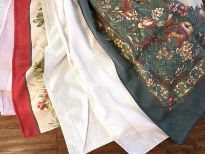 Assorted vintage table linens, including a white tablecloth, a holiday-themed runner with red borders, and a green floral patterned cloth.