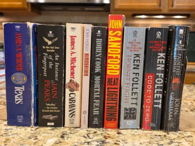 Collection of vintage paperback novels including titles by James A. Michener, Ken Follett, and John Grisham, showing some shelf wear on...