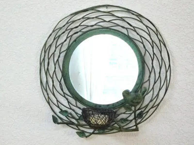 Decorative round wall mirror with a green-patinated metal wire frame designed to look like a bird's nest, featuring a small bird...