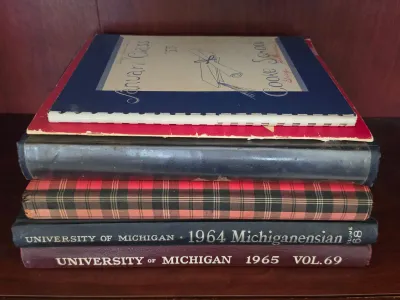 Stack of vintage yearbooks and school memorabilia, including University of Michigan Michiganensian volumes from 1964 and 1965, showing...