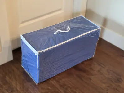Blue fabric storage chest or organizer with a clear plastic zippered cover and a white rope handle, shown sitting on a dark wood floor.