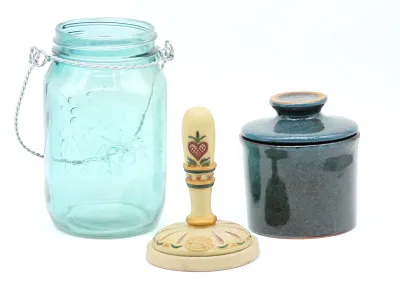 Vintage kitchenware trio featuring a blue glass Ball jar with a wire handle, a hand-painted wooden butter stamp with a heart motif, and a...