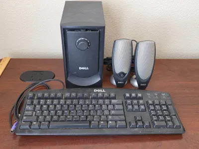 Vintage Dell computer accessories, including a black PS/2 keyboard, a subwoofer, and a pair of desktop speakers, showing some dust and...