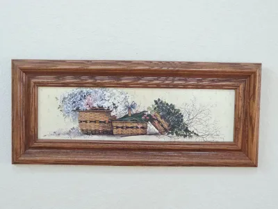 Framed country-style print depicting woven baskets filled with flowers and greenery, housed in a wide oak frame.