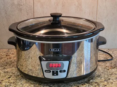 Bella digital slow cooker with a polished stainless steel exterior, black ceramic insert, and glass lid.
