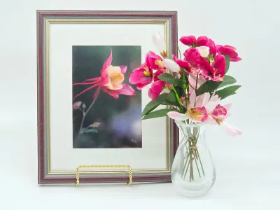 Framed photograph of a pink columbine flower on a gold display stand, paired with a small clear glass vase holding artificial pink flowers.