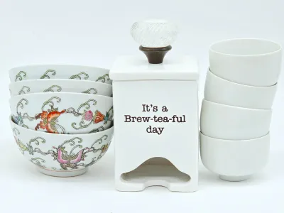 Ceramic tea set featuring a white tea bag dispenser reading "It's a Brew-tea-ful day," flanked by a stack of four...