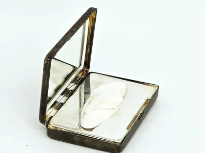 Vintage silver-plated makeup compact featuring an interior mirror and a powder puff, showing some tarnish and wear consistent with age.