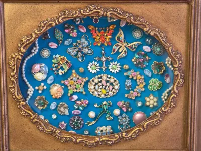 Framed vintage costume jewelry display featuring an assortment of colorful brooches, pins, and faux pearls arranged on a blue velvet...