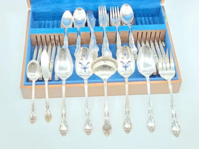 Vintage silver-plated flatware set with ornate handles, displayed in a blue felt-lined wooden storage chest.