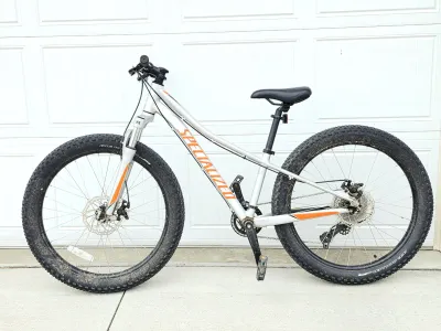 Specialized Riprock youth mountain bike with a silver frame, orange accents, front suspension, and wide knobby tires.