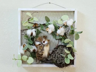 Rustic shadow box wall hanging featuring a faux owl, bird's nest, cotton bolls, and eucalyptus leaves arranged on a chicken wire...