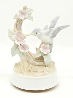 Vintage porcelain music box figurine featuring a hummingbird hovering near pink dogwood blossoms, rendered in soft pastel tones with a...