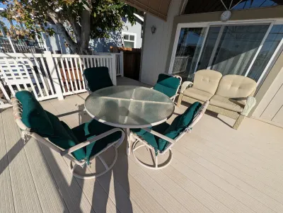 Outdoor patio dining set featuring a round glass-top table and four swivel chairs with green cushions, alongside a beige wicker loveseat.