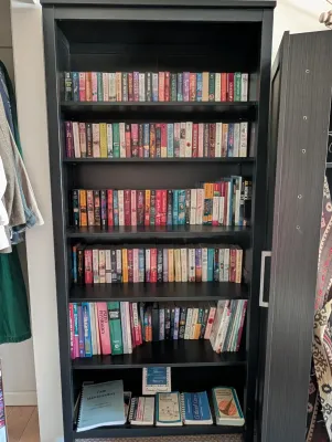 Tall black wooden bookcase with six shelves, filled with a large collection of paperback novels and reference books.  Bookshelf also for...