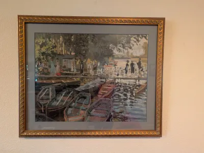 Framed impressionist-style print depicting a lakeside scene with figures and rowboats, presented in an ornate gold-finished frame with a...