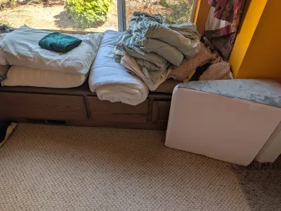 Assorted bedding including pillows, sheets, and a comforter stacked on a wooden window bench, alongside a foam wedge pillow.