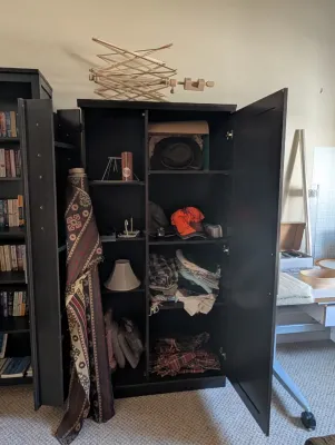 Dark brown wooden storage cabinet with double doors, featuring adjustable shelves filled with various textiles, hats, and household...