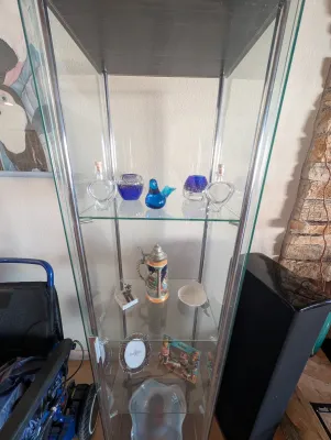 Modern glass and chrome curio cabinet displaying various collectibles, including art glass, a German beer stein, and small picture frames.