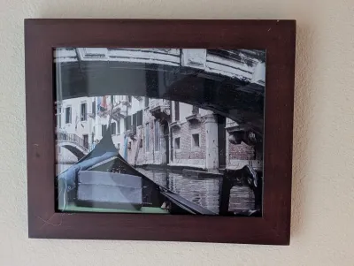 Framed photograph depicting a Venetian canal scene with a gondola passing under a bridge, housed in a simple dark wood frame.