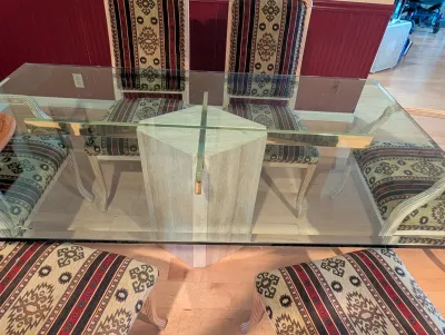 Postmodern dining table featuring a thick rectangular glass top supported by a travertine stone pedestal base with brass accents,...