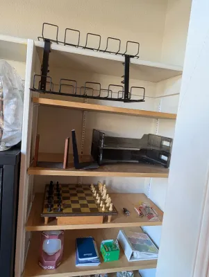 Wooden shelving unit holding various household items, including a chess set, office organizers, and a wire storage rack.